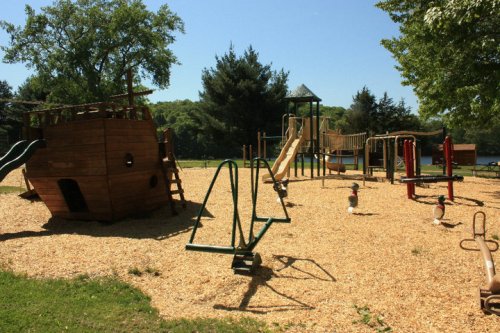 A playground featuring a wooden ship structure, slides, swings, and various play equipment on a sunny day.