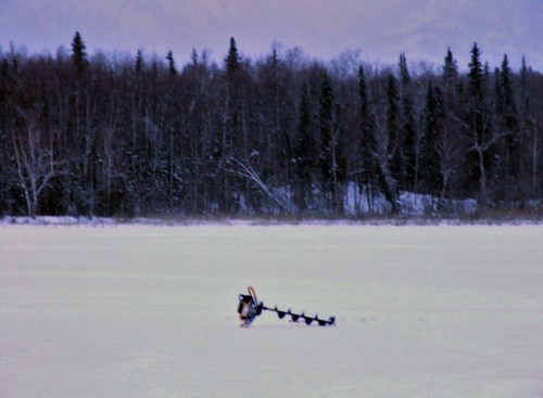 A person using an auger to drill through ice on a frozen lake, surrounded by a forest of trees in the background.