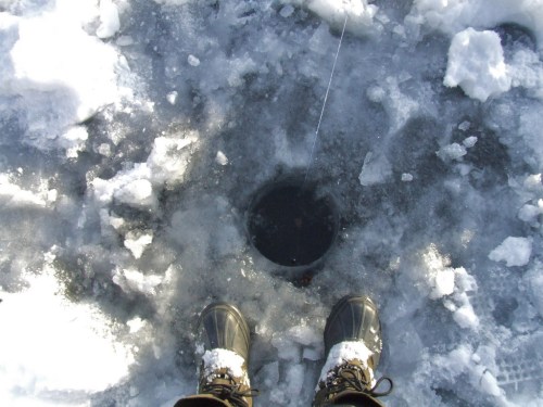 Person's feet in winter boots standing over a hole in the ice, surrounded by snow and ice shards.