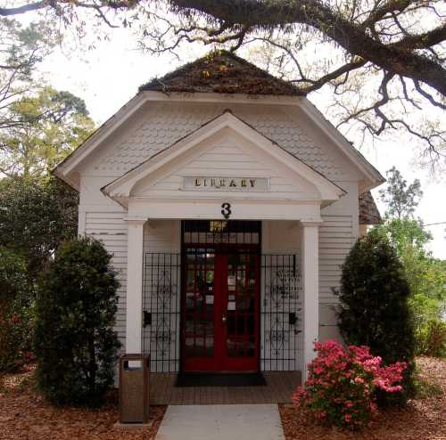 A small, charming library building with a peaked roof, red doors, and surrounded by greenery and flowers.