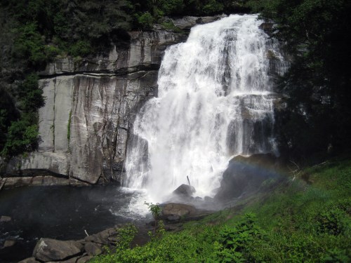 A cascading waterfall flows over rocky cliffs into a serene pool, surrounded by lush greenery and sunlight.