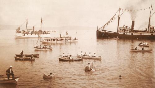Historic sepia-toned image of boats and a steamship on calm waters, with people enjoying a day out on the water.