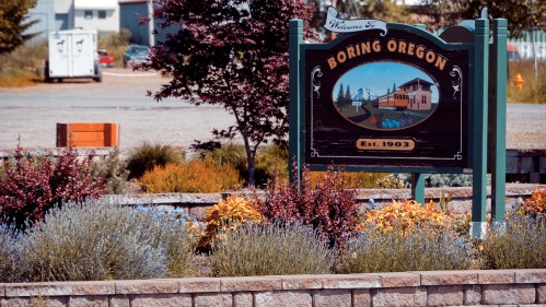 Welcome sign for Boring, Oregon, established in 1903, surrounded by colorful plants and a clear blue sky.