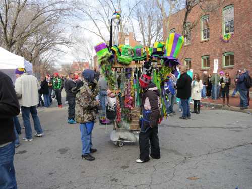 A street vendor cart filled with colorful hats and beads, surrounded by people at a festive outdoor event.