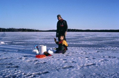 A child and an adult stand on a snowy landscape, with ice and trees in the background, engaged in an outdoor activity.