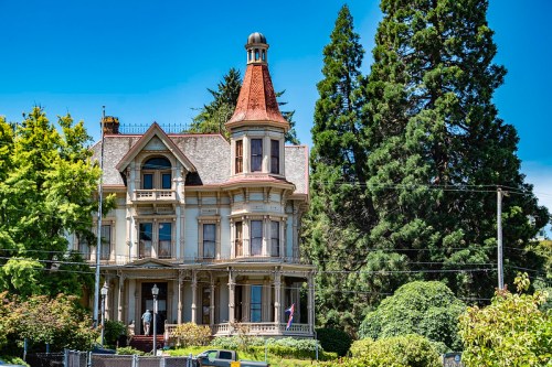A grand Victorian-style mansion with a turret, surrounded by lush greenery and tall trees under a clear blue sky.
