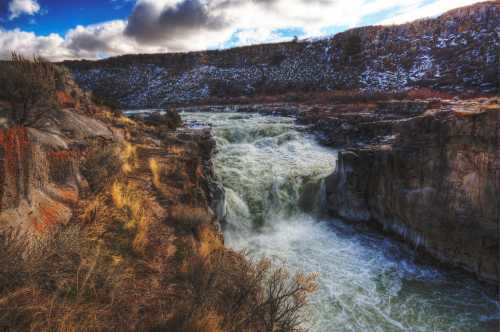 A scenic view of a rushing river cascading over rocky cliffs, surrounded by rugged terrain and cloudy skies.