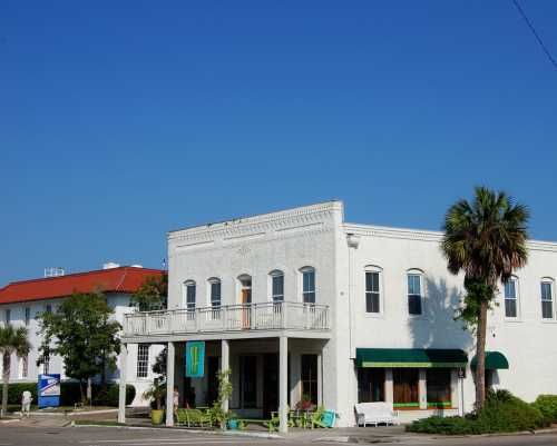 A white building with a balcony, surrounded by greenery, under a clear blue sky. Palm trees are visible nearby.