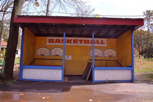 Abandoned basketball court structure with faded backboards and a sign, surrounded by trees and fallen leaves.