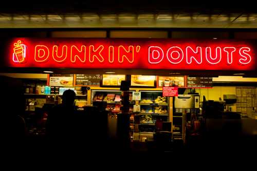 Bright neon sign of Dunkin' Donuts with a silhouette of a person at the counter, showcasing a variety of menu items.