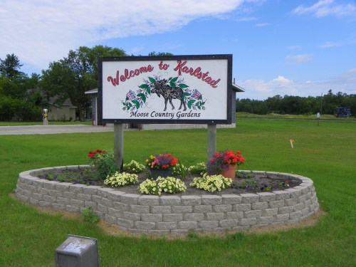 Welcome sign for Keshad featuring flowers and a moose design, set in a landscaped area under a blue sky.