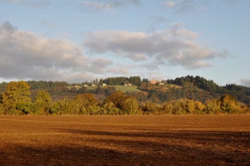 A scenic view of a plowed field with rolling hills and a small village in the background under a partly cloudy sky.