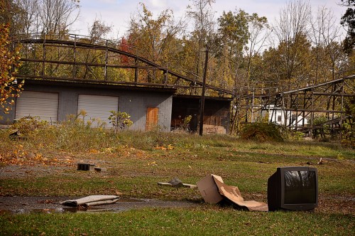 An abandoned amusement park with a dilapidated roller coaster, overgrown grass, and a discarded old television.