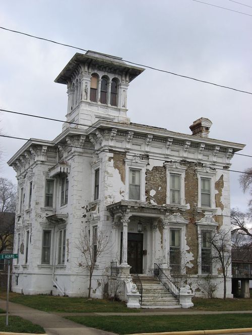 A dilapidated Victorian-style house with peeling white paint and ornate architectural details under a cloudy sky.