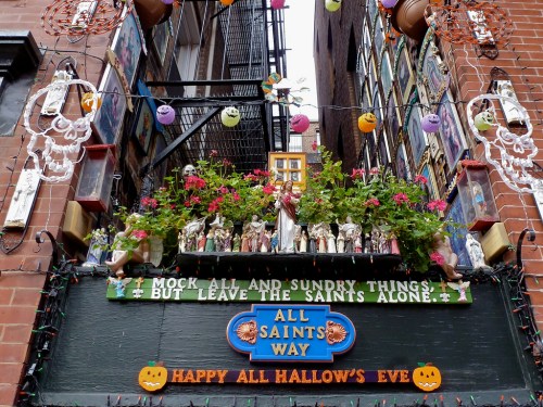 Colorful decorations and flowers adorn a sign reading "All Saints Way" with Halloween-themed elements above a brick wall.