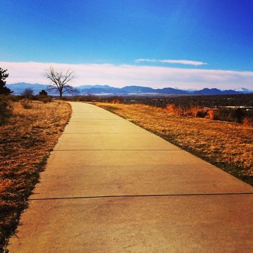 A paved path winds through a grassy area, leading towards distant mountains under a clear blue sky.
