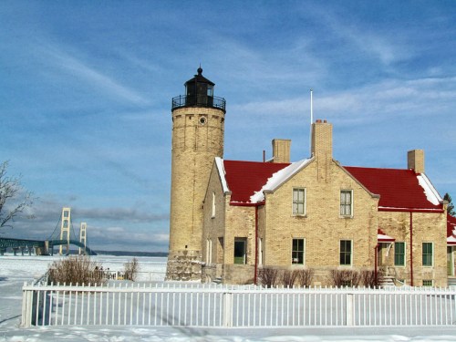 A historic lighthouse with a red roof, surrounded by snow, near a bridge under a clear blue sky.