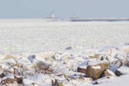 A snowy landscape with ice-covered rocks in the foreground and distant lighthouses on a frozen sea.