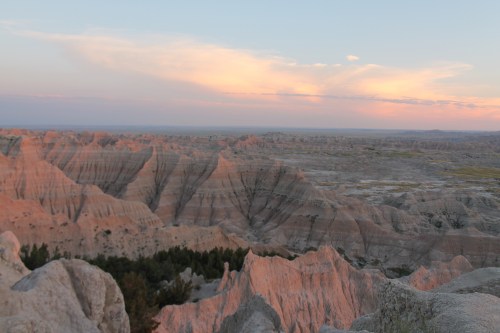 A panoramic view of rugged, colorful rock formations under a pastel sky at sunset in a vast landscape.