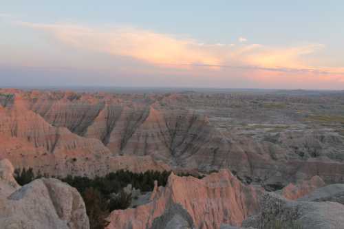 A panoramic view of rugged, colorful rock formations under a pastel sky at sunset in a vast landscape.