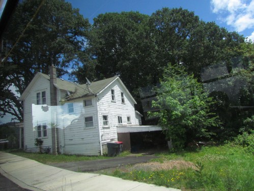 A white, two-story house with a garage, surrounded by greenery and trees under a blue sky.