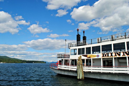 A white steamboat docked by the shore, with blue skies and fluffy clouds above a calm lake.