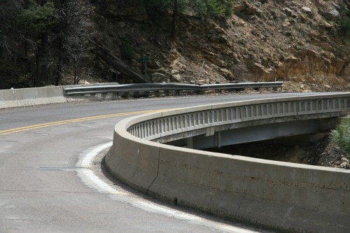 A curved road with a concrete barrier, surrounded by rocky terrain and greenery.