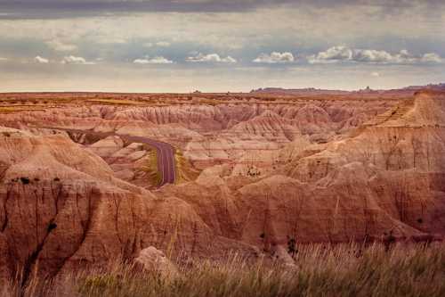 A winding road cuts through rugged, colorful badlands under a cloudy sky.