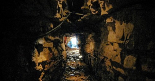 A dimly lit tunnel with rough stone walls, leading to a bright exit in the distance.