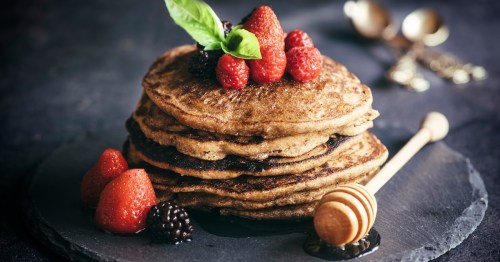 close up of a stack of buckwheat pancakes topped with strawberries and blackberries on a black plate next to a honey comb wand