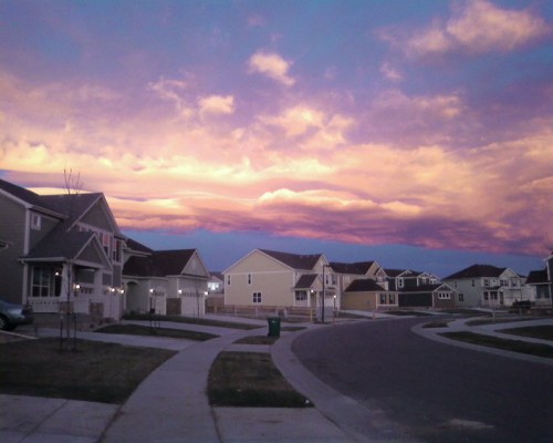 A suburban neighborhood at dusk, featuring houses and a colorful sunset sky with vibrant clouds.
