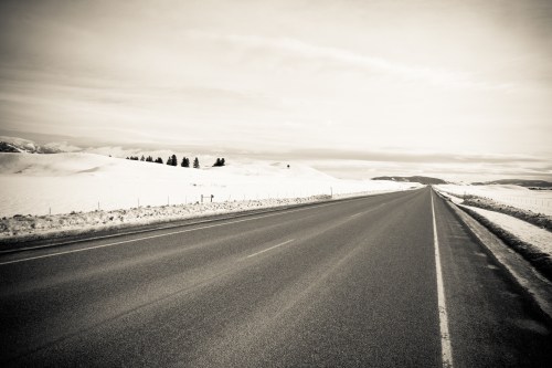A desolate, snow-covered road stretches into the distance, flanked by white hills and a cloudy sky.