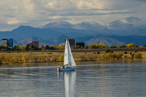 A sailboat glides across a calm lake with mountains and buildings in the background under a cloudy sky.