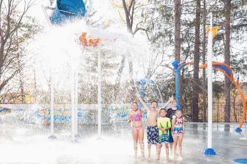 Five children play joyfully in a splash pad, water spraying around them on a sunny day.
