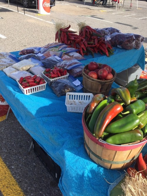 A vibrant farmers market table displaying various fresh vegetables, including peppers, apples, and bags of spices.