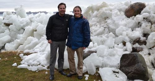 Two men stand smiling in front of a large pile of ice and rocks, with a cloudy sky in the background.