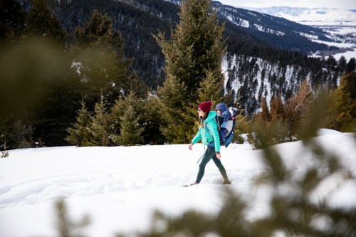 A person in a green jacket and red hat walks through snowy terrain, surrounded by evergreen trees and mountains.