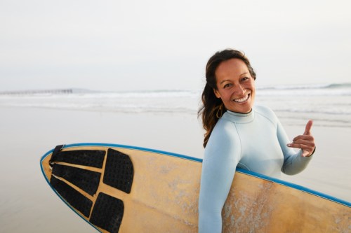 A smiling woman in a wetsuit holds a surfboard, making a shaka sign on a beach with waves in the background.