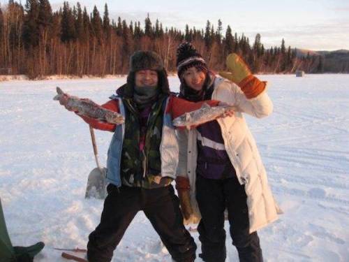 Two people in winter clothing hold up large fish while standing on a snowy landscape with trees in the background.