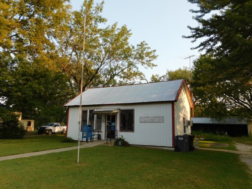A small white building with a red roof, surrounded by trees and grass, featuring a mailbox and a flagpole in front.