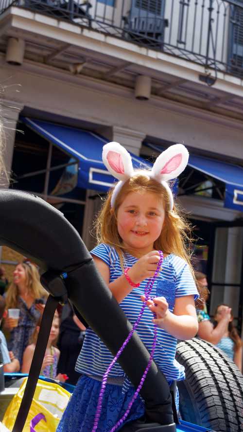 A young girl with bunny ears smiles while holding beads at a festive outdoor event.