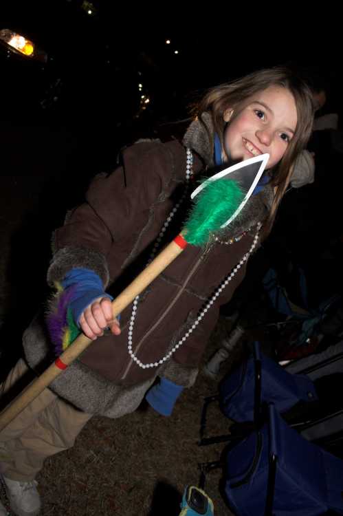 A smiling child in a brown coat holds a colorful spear, surrounded by a nighttime outdoor setting.