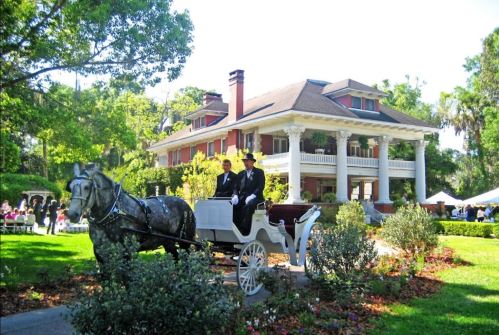 A horse-drawn carriage in front of a grand mansion, surrounded by lush greenery and people enjoying an outdoor event.