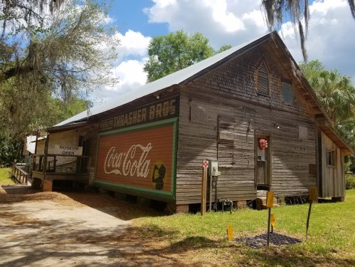 A rustic wooden building with a Coca-Cola sign, surrounded by greenery and trees under a partly cloudy sky.