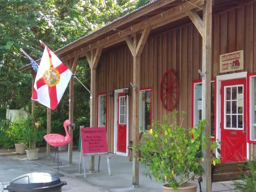 A wooden building with red doors, a Florida flag, a pink flamingo, and a sign in front surrounded by greenery.