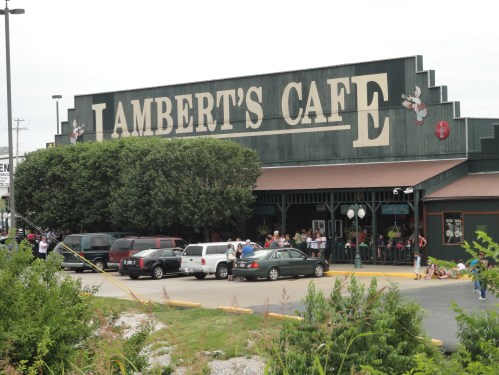 A large, rustic building with "Lambert's Cafe" sign, featuring a line of people waiting outside.