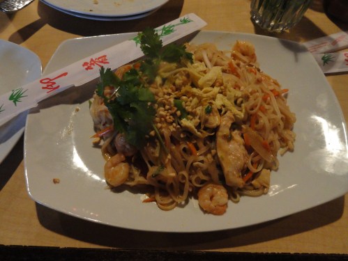 A plate of pad thai with shrimp, chicken, vegetables, peanuts, and cilantro, served with chopsticks on the side.