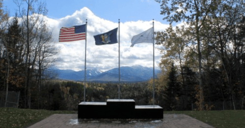 Three flags stand on a stone pedestal with mountains and a cloudy sky in the background, surrounded by trees.