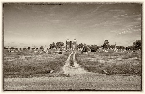 A gravel path leads through a cemetery towards a distant church, surrounded by gravestones and greenery.