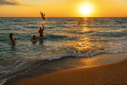 A family enjoys a sunset at the beach, with one person playfully tossing a child into the air above the waves.